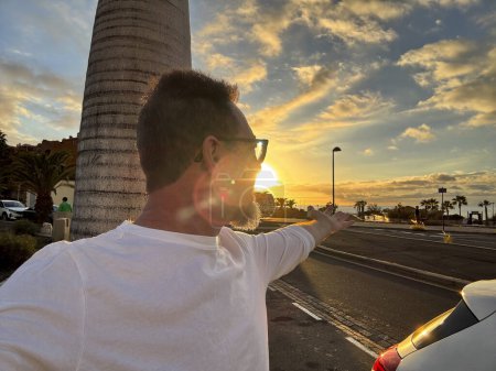 Foto de Happy man taking portrait selfie picture against a colorful sunset on the street with car and roads. Travel and summer vacation destination concept lifestyle and outdoor leisure activity. Happiness - Imagen libre de derechos