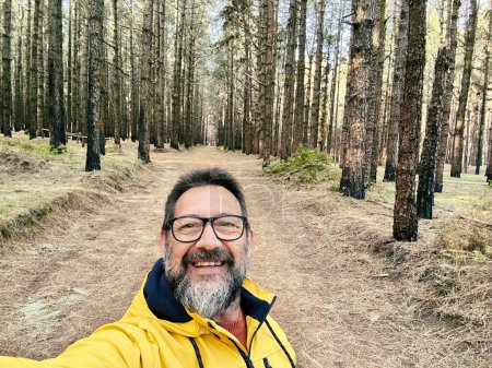 Photo for Cheerful adult happy man taking selfie picture in the woods enjoying outdoors leisure activity in national forest park alone. Adventure and lifestyle people. High trees in background. Sustainability - Royalty Free Image