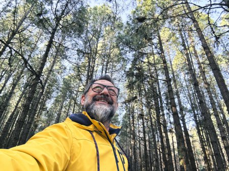 Foto de Alegre adulto feliz hombre tomando foto selfie en el bosque disfrutando de la actividad de ocio al aire libre en el parque forestal nacional solo. Aventura y estilo de vida. Árboles altos en el fondo. Sostenibilidad - Imagen libre de derechos