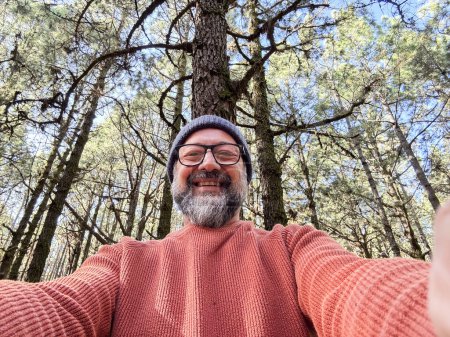 Foto de Alegre adulto feliz hombre tomando foto selfie en el bosque disfrutando de la actividad de ocio al aire libre en el parque nacional forestall solo. Aventura y estilo de vida. Árboles altos en el fondo. Sostenibilidad - Imagen libre de derechos
