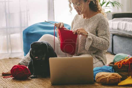 Foto de Mujer sentada en el suelo tejiendo una bola de lana roja con su perrito negro junto a ella. aprender viendo tutoriales en el portátil los domingos por la tarde. Estilo de vida y ocio concep - Imagen libre de derechos