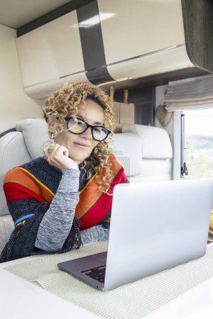 Foto de Vivir y trabajar dentro de la caravana es un estilo de vida digital libre de nómadas. Una mujer sentada en una caravana disfruta de relajarse y conectarse a su computadora portátil. bosque en el fondo fuera de la ventana - Imagen libre de derechos