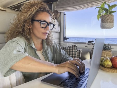 Foto de Mujer caucásica rizada con gafas trabajando en su portátil en su caravana. vacaciones en casa sobre ruedas y aventura primaveral en la playa. - Imagen libre de derechos