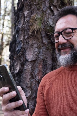 Téléchargez les photos : Homme utilisant le téléphone dans un bois forestier profitant de la technologie et des loisirs de plein air. Homme moderne parlant au cellulaire avec des arbres en arrière-plan. Personnes et communication sur les voyages - en image libre de droit