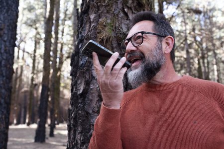 Foto de Hombre usando el teléfono en un bosque de madera disfrutando de la tecnología y la actividad de ocio al aire libre. Hombre moderno hablando en celular con árboles en el fondo. Personas y comunicación sobre viajes - Imagen libre de derechos
