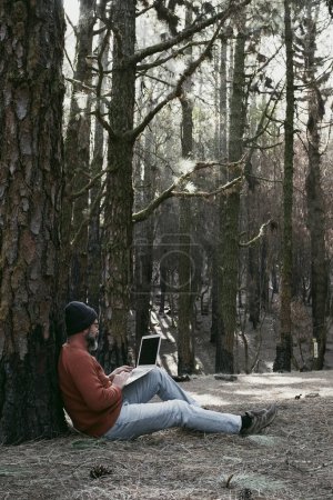 Photo for Man using laptop in a forest wood enjoying technology and outdoor leisure activity. Modern man speaking at cellular with trees in background. People and communication on travel - Royalty Free Image