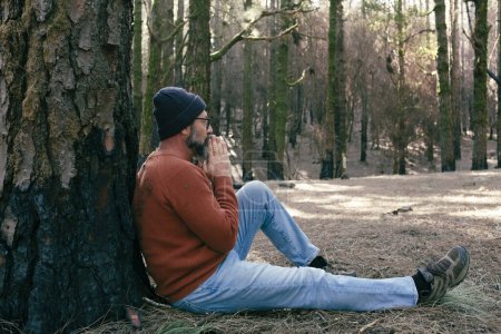 Téléchargez les photos : Homme ayant du temps de détente assis sur le sol au milieu de forêts verdoyantes et activités de loisirs de détente en plein air. Les gens et l'amour sentiment avec le parc naturel. Bien-être mode de vie alternatif - en image libre de droit