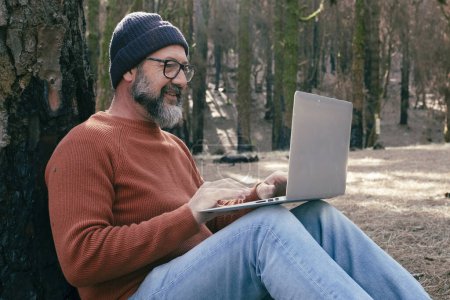 Foto de Hombre usando el ordenador portátil en un bosque de madera disfrutando de la tecnología y la actividad de ocio al aire libre. Hombre moderno hablando en celular con árboles en el fondo. Personas y comunicación sobre viajes - Imagen libre de derechos
