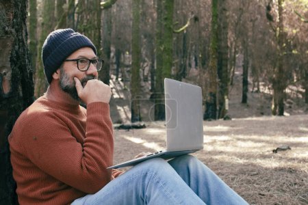 Téléchargez les photos : Homme utilisant un ordinateur portable dans un bois forestier profiter de la technologie et des loisirs de plein air. Homme moderne parlant au cellulaire avec des arbres en arrière-plan. Personnes et communication sur les voyages - en image libre de droit
