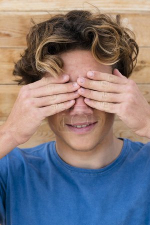 Photo for Young caucasian teenager hide his eyes and smile - youthful and teen generation z concept with happy and nice male boy with blue t-shirt and wooden background - Royalty Free Image