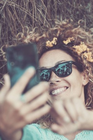 Téléchargez les photos : Beau portrait de heureuse femme caucasienne gaie faire selfie image avec téléphone moderne - feuille brune et la nature autour de sa tête et sur ses cheveux - profiter de la nature et concept de forêt - en image libre de droit
