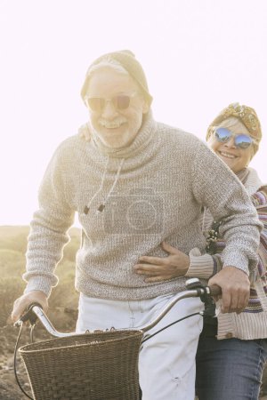 Téléchargez les photos : Couple de personnes âgées actives gaies et heureuses retraités aiment ensemble faire du vélo dans une activité de loisirs de plein air avec soleil dans le rétroéclairage concept de vieillesse et de plaisir joyeux sans limite - en image libre de droit