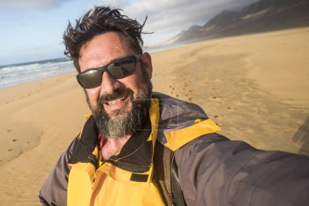 Foto de Hombre en la naturaleza salvaje en la playa haciendo foto selfie - montañas y nadie más en el fondo - explorar y viajar estilo de vida para las personas y la vida alternativa y vacaciones con mochila - Imagen libre de derechos