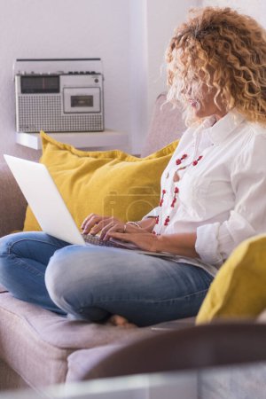 Téléchargez les photos : Femme adulte à la maison dans le travail intelligent avec ordinateur portable s'asseoir sur le canapé écriture et profiter de la technologie et de l'appareil de communication - belles femmes dans l'activité de travail de vidéoconférence - en image libre de droit