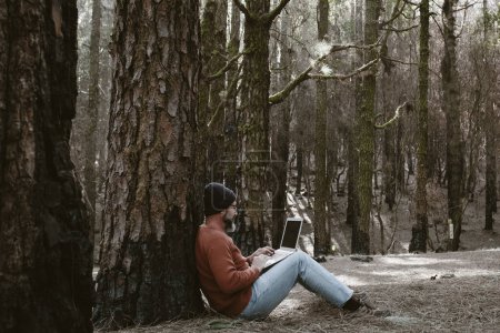 Photo for Man using laptop in a forest wood enjoying technology and outdoor leisure activity. Modern man speaking at cellular with trees in background. People and communication on travel - Royalty Free Image