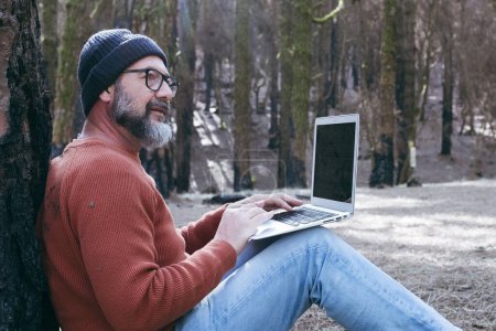 Téléchargez les photos : Homme utilisant un ordinateur portable dans un bois forestier profiter de la technologie et des loisirs de plein air. Homme moderne parlant au cellulaire avec des arbres en arrière-plan. Personnes et communication sur les voyages - en image libre de droit
