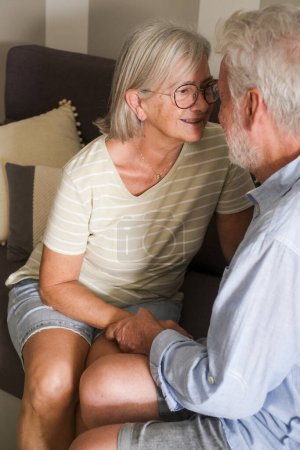 Love and care with mature senior couple at home looking each other sitting on the sofa. Retired people indoor leisure activity and love. Elderly lifestyle. Caring and talking people 80 years old