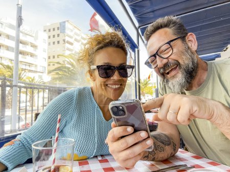 Photo for Smiling couple enjoying time at the restaurant looking at the phone and talking outdoors having fun together. Consumerism, food, lifestyle concept - Royalty Free Image