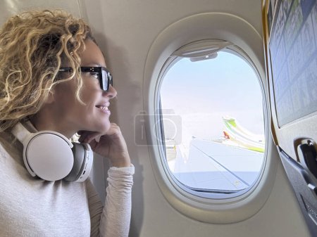 Foto de Vista lateral del pasajero femenino dentro del vuelo del avión mirando fuera de la ventana el ala y el cielo azul sonriendo y disfrutando del viaje escuchando música en los auriculares. Viajes y vacaciones gente feliz - Imagen libre de derechos