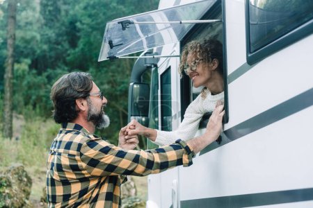 Couple man and woman enjoy camper van travel destination, one outside the van and the female inside at the window with green forest park nature in background and around. Vanlife and vacation people