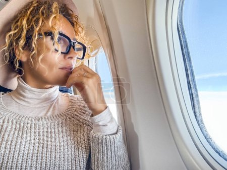 Foto de Atractiva viajera femenina con gafas mirando fuera de la ventana sentada dentro del vuelo del avión. Concepto de personas y transporte aéreo. Destino de vacaciones. Mujer adulta en viaje - Imagen libre de derechos