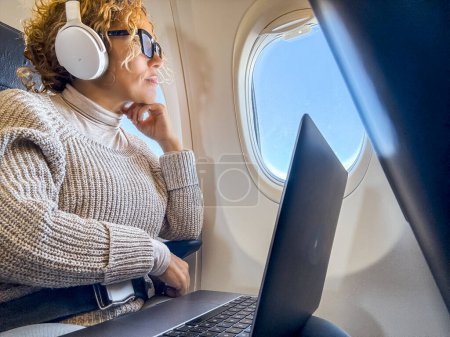 Téléchargez les photos : Passagère assise à l'intérieur du vol d'avion profitant du voyage pour écouter de la musique ou regarder un film sur ordinateur portable. Personnes voyageant pour des vacances d'affaires ou de vacances seules. Transport aérien - en image libre de droit