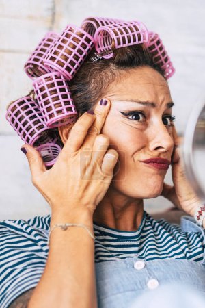 Foto de Preocupada mujer caucásica adulta comprobar las arrugas y el efecto de envejecimiento en su piel de la cara hermosa mujer divertida con maquillaje y rizadores en el pelo en casa en la preparación de la actividad - Imagen libre de derechos