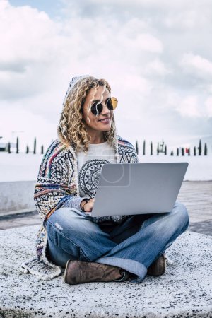 Téléchargez les photos : Smart travail et numérique nomade gens concept de style de vie avec gai heureux et libre femme caucasienne travaillant en plein air avec ordinateur portable sur ses jambes - les gens modernes et la connexion technologique - en image libre de droit
