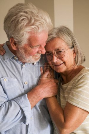 Photo for Love at senior age between a mature man and old lady at home hugging and protecting each other with romantic expression. Portrait of retired people having care and enjoying elderly lifestyle together - Royalty Free Image