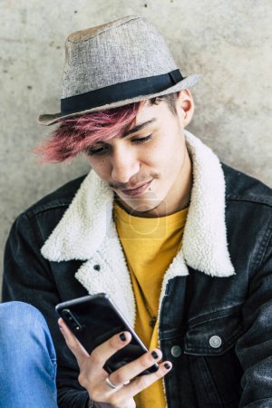 Photo for Young boy teenager alone sitting on the ground using mobile phone app to communicate and surfing the net on social media. Teenager scrolling phone outdoor. Alternative hairstyle and lifestyle. Modern - Royalty Free Image