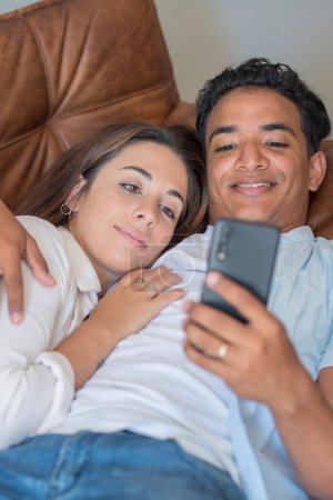 Téléchargez les photos : Jeune couple ayant se détendre ensemble en utilisant le téléphone portable couché sur le canapé dans l'activité de loisirs de détente intérieure ensemble. Garçon noir et fille caucasienne dans l'amour et la relation, Les gens intimes profiter - en image libre de droit
