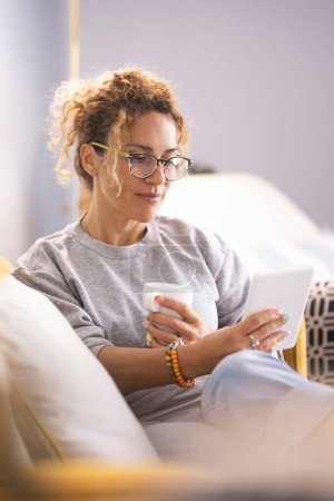 Modern lady having relax at home sitting on couch and using reader tablet to read an online ebook. Woman using technology indoor leisure activity alone enjoying relaxation on couch. Indoor life