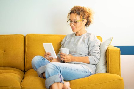Téléchargez les photos : Smart casual femme d'âge moyen relaxant à la maison assis sur un canapé jaune et en utilisant le lecteur pour lire un ebook profiter d'une activité de loisirs en intérieur seul. Style de vie du dimanche. Personnes et technologie en ligne - en image libre de droit
