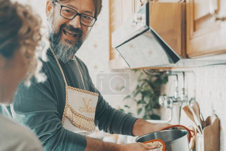 Foto de Pareja real cocinando juntos en casa en la cocina. Un hombre sonríe a una mujer y usa ollas para preparar el almuerzo. Marido y esposa viviendo en apartamento. Personas maduras en interior actividad de ocio felicidad - Imagen libre de derechos