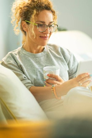 woman reading ebook on electronic reader tablet sitting comfortably on the sofa at home in indoor leisure activity. People with reader enjoying relax indoor on couch. Happy female people inside