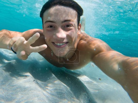 Young boy enjoy summer holiday vacation taking selfie underwater swimming at the sea with sand ground and blue water. Happy tourist swim in crystal clean ocean water doing victory gesture with hands