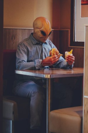 Téléchargez les photos : Homme avec masque extraterrestre manger seul dans un magasin de restauration rapide hamburger de mauvaise nourriture et frites. Ufo sont parmi nous vivant comme concept humain drôle d'image. Invasion et diversité personnes loisirs intérieurs - en image libre de droit