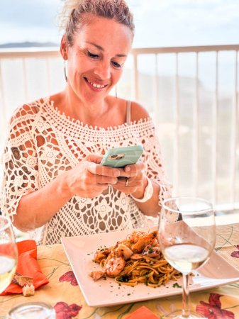 Foto de Mujer adulta bonita tomando la foto de su comida antes de comer. Pasta de espaguetis con pescado. Las mujeres modernas fotografian el almuerzo usando el teléfono móvil para compartir contenido en línea estilo de vida creador - Imagen libre de derechos