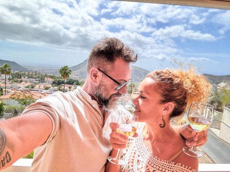 Téléchargez les photos : Heureux couple adulte prenant selfie photo à la maison en terrasse avec vue sur la ville et les montagnes en plein air. Les gens aiment les vacances et les loisirs ensemble, souriant et buvant du vin. Joyeuses vacances des gens - en image libre de droit