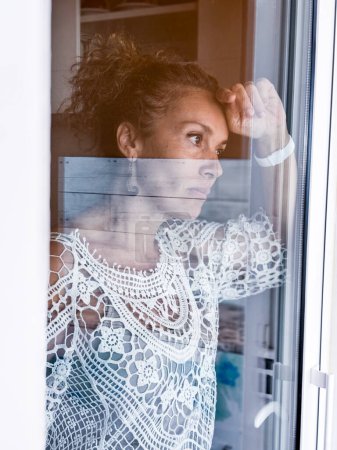 Photo for Sad adult woman inside home looking outside through the door window glass. Concept of mental burnout and depression. Lonely lady sad expression portrait. Breakup relationship love divorced lady - Royalty Free Image