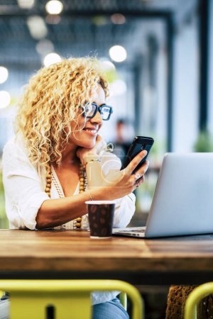 Photo for Happy  adult woman using phone and laptop at the desk in cafe with free wireless connection. Modern businesswoman working online in coworking space. Smart people with online job indoor activity - Royalty Free Image
