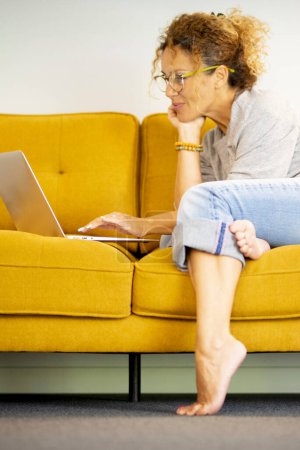Photo for Happy woman freelancer work on computer typing texting from home, technology concept. One female people using laptop on the yellow sofa and smiling. Surfing web activity. Side view. Interior. - Royalty Free Image