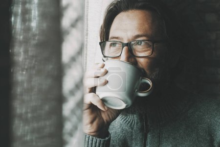 Foto de Hombre con expresión reflexiva y serena en la cara, bebiendo café o té solo en casa mirando fuera de la ventana. Hombres maduros solos. Concepto de estilo de vida saludable y consciente. Usar gafas - Imagen libre de derechos