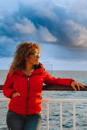 Téléchargez les photos : Femme profiter et admirer coucher de soleil ciel de couleur debout à l'extérieur du ferry sur le quai. Veste rouge. Vacances mode de vie de transport vacances - en image libre de droit