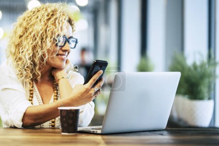 Photo for Happy y adult woman using phone and laptop at the desk in cafe with free wireless connection. Modern woman working online in coworking space. Smart people with online job indoor activity - Royalty Free Image