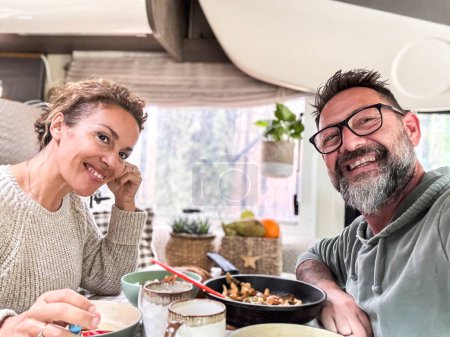 Photo for Happy couple eating together inside a camper van motorhome living the vanlife and travel adventure holiday vacation. Nomadic life. Cheerful man and woman taking selfie and smiling at the camera - Royalty Free Image