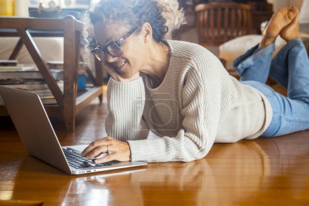 Téléchargez les photos : Femme détendue à l'aide d'un ordinateur à la maison posé sur le sol en bois dans un appartement moderne et un mode de vie serein. Recherche sur le web. Loisirs intérieurs avec ordinateur portable et technologie. Envoi de chat par e-mail - en image libre de droit