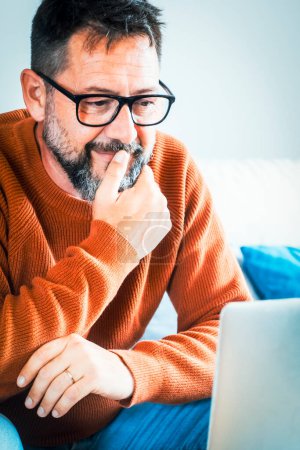 Foto de Adulto freelance hombre mirando la pantalla del ordenador portátil pensando y tocando la barbilla. Usando gafas maduras masculinas. Trabajo en el hogar inteligente trabajo remoto concepto de negocio estilo de vida. Trabajador moderno sentado en el sofá - Imagen libre de derechos