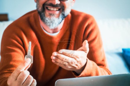 Photo for One adult man showing home key sitting on sofa with open laptop in front. Real estate renting vacation holiday properties working form home sitting on sofa. New apartment owner showing keys on camera - Royalty Free Image