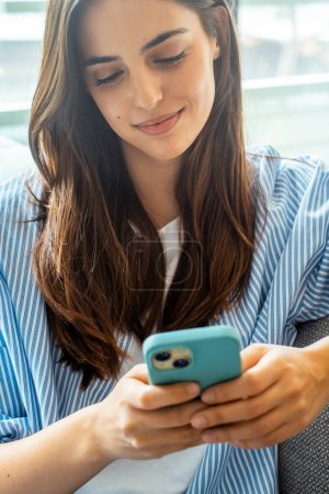 Happy girl checking social media while holding smartphone at home. Smiling young brunette woman using mobile phone app to play games, shop online, order delivery while relaxing on sofa